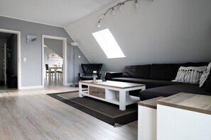 Um espaço de estar moderno em estilo loft, com teto inclinado, sofá de couro preto, mesa de centro branca e um espaço de refeições ao fundo.