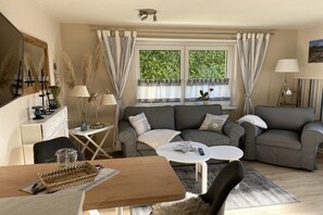 Ein Wohnzimmer mit einer Sofagarnitur, einem Couchtisch, einem Esstisch und einem Fenster mit Vorhängen.