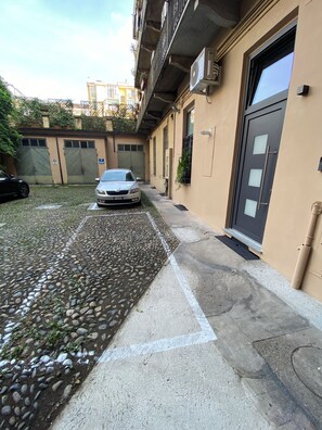 A parking area with a paved surface and a gravel section, a silver car parked, and a building with a dark door and window. - Appartamenti Madama, new apartments with private parking (Torino)