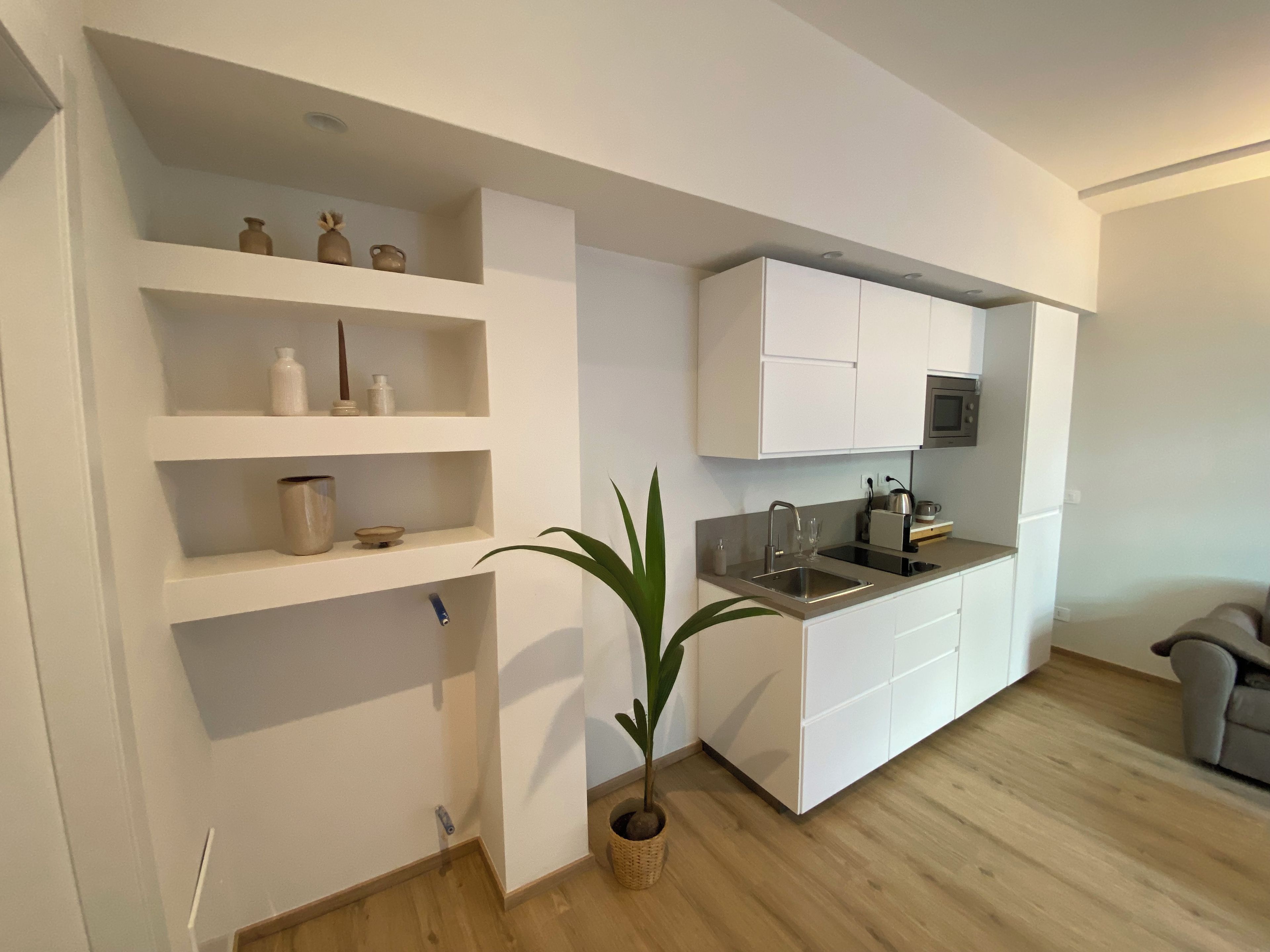 A modern kitchen with white cabinetry, a built-in microwave, and a sink. There are shelves with various kitchen items and a potted plant.