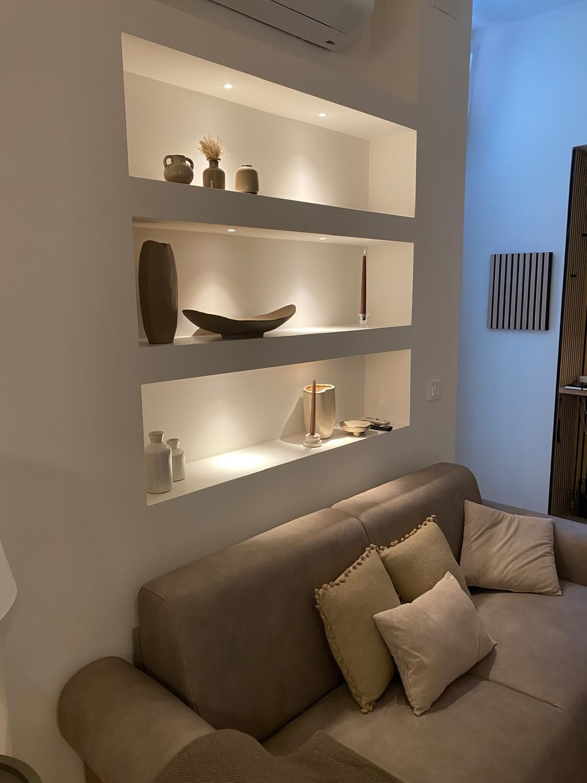 A modern living room with a beige sofa, a wall-mounted shelf with decorative items, and a minimalist decor style.