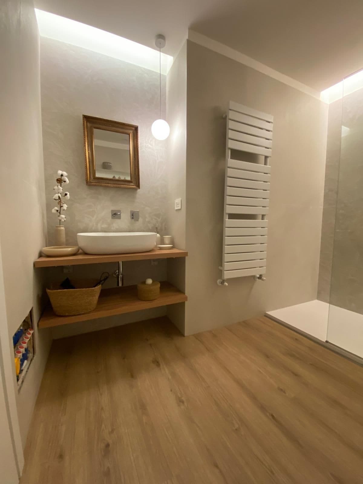 Une salle de bain avec un plancher en bois, un lavabo blanc avec un miroir au-dessus, un porte-serviettes et un espace douche.