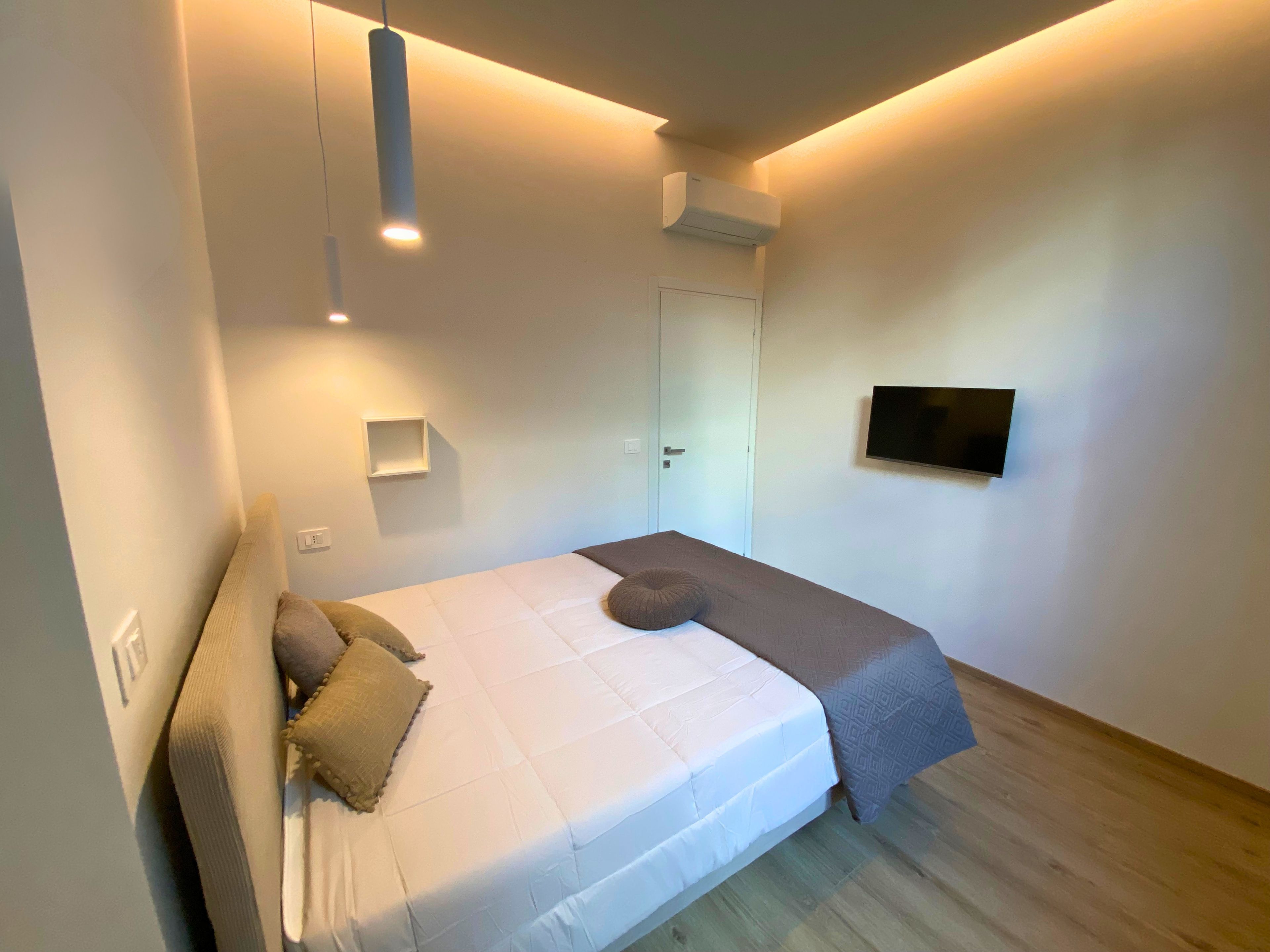 Un dormitorio con cama, mesita de noche, televisor en la pared y una puerta.