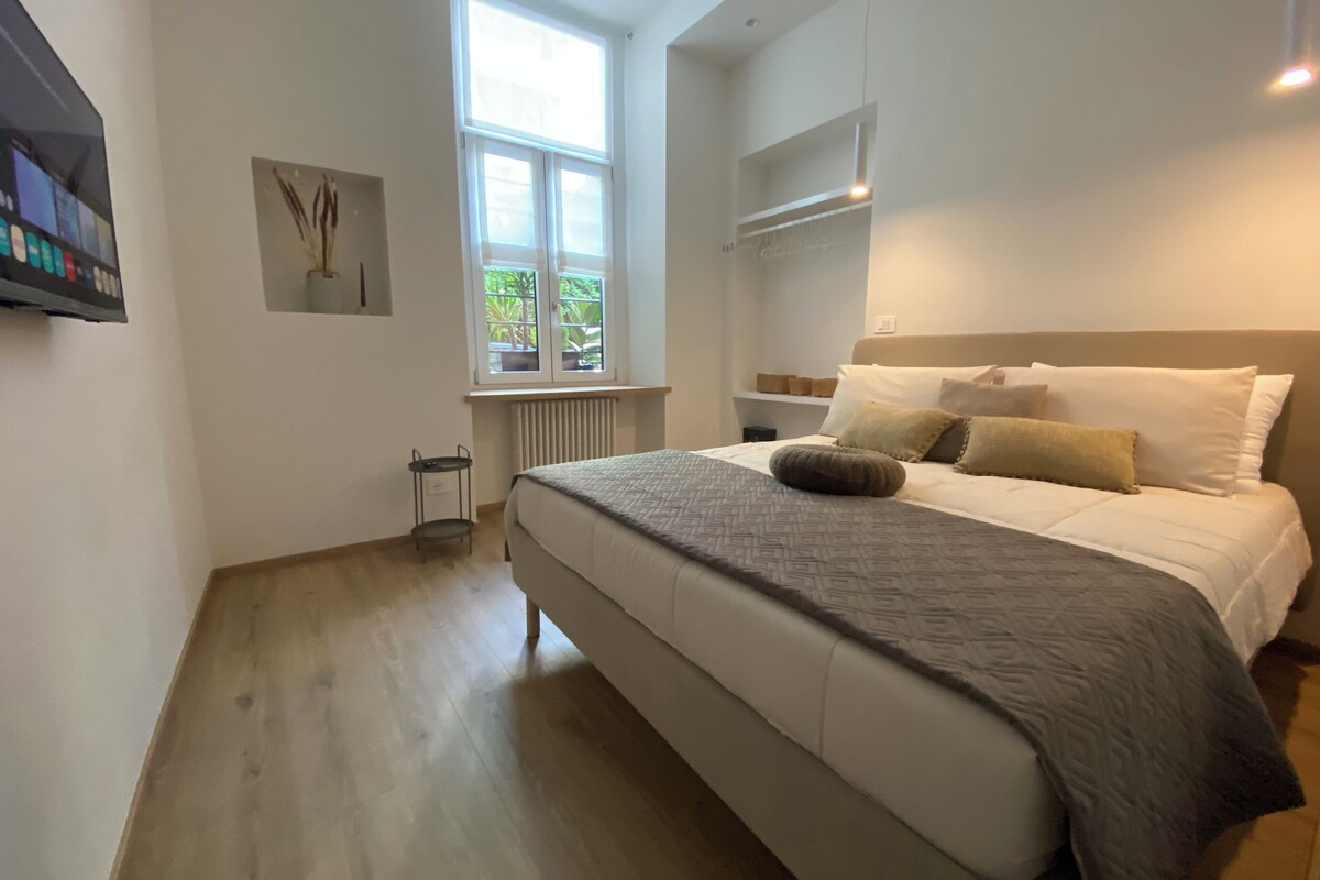 A bedroom with a large bed, a window with a view of greenery, a radiator, and a wall-mounted shelf.