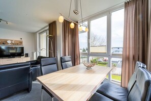 Een moderne woonkamer met een houten eettafel, zwarte leren banken, een flatscreen televisie en een groot raam met gordijnen.