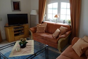 A living room with a sofa, a television on a wooden stand, a glass coffee table with a plant, and a window with curtains. - A holiday with flair - Sternberg (Sternberg)