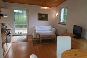 Ein gemütliches Wohnzimmer mit einem weißen Sofa, einem Holztisch, einem Fernseher und einem Fenster mit Blick auf Grünflächen.