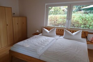 Ein Schlafzimmer mit einem Doppelbett, einem Holzschrank, einem Nachttisch, einer Lampe und einem Fenster mit Blick auf Grünflächen.