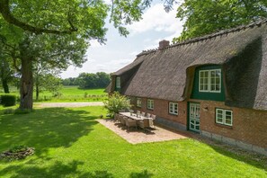 Ein Haus mit einem Strohdach und einer Ziegelaußenwand, eine Terrasse mit Tisch und Stühlen sowie ein gepflegter Rasen.