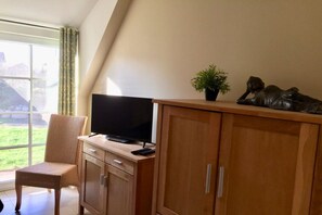 Ein Zimmer mit Fernseher auf einer Holzkommode, einem Sessel und einer Topfpflanze.