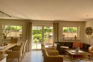 Ein großzügiger Wohnbereich mit Esstisch, Stühlen, einer Couch und einem Couchtisch. Große Fenster bieten einen Blick ins Freie.