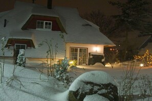 Ein mit Schnee bedecktes Haus mit einem beleuchteten Fenster, einer sichtbaren Tür und einem Schneebedeckten Felsen im Vordergrund.