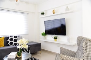 Uma sala de estar moderna com TV de tela plana na parede, um sofá cinza, uma poltrona estampada e uma mesa de centro com livros e um vaso de flores.