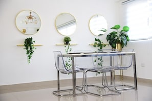 Uma sala de jantar com uma mesa de vidro transparente e cadeiras, três espelhos com moldura dourada e plantas em vasos sobre prateleiras.