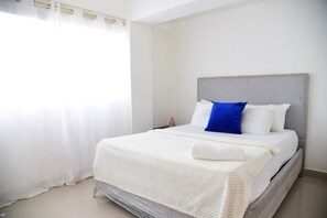 Cama bem arrumada com cabeceira cinza, roupa de cama branca e almofada azul de destaque.