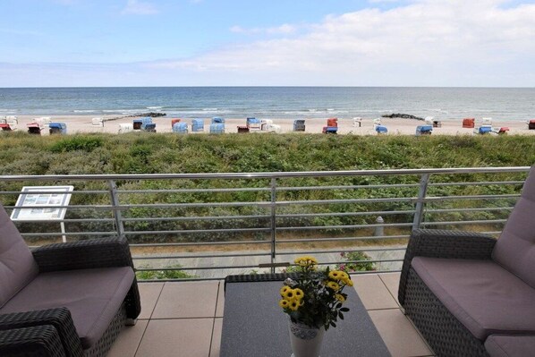 Ein Balkon mit Korbmöbeln, einem Tisch mit geliler Blumenarragem und Blick auf den Strand mit Liegestühlen.