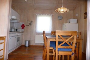 A compact kitchen with wooden cabinets, a sink, a stove, and a refrigerator. There is a dining table with chairs and a window with a curtain. - Holiday home with 2 bedrooms (Otterndorf)