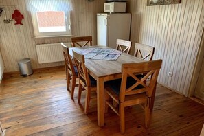 A dining area with a wooden table and chairs, a window with a view, a refrigerator, and a trash bin. - Holiday home with 2 bedrooms (Otterndorf)