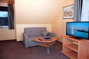 Ein Zimmer mit grauem Sofa, einem kleinen Holztisch, einem Fernseher auf einem Schrank und einem Fenster mit Vorhängen.