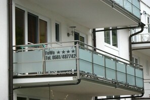 Un balcone con ringhiera in vetro, un cartello con i recapiti e una finestra con persiane.