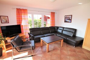 Ein Wohnzimmer mit einem schwarzen Ledersofa, einem Holztisch, einem schwarzen Sessel, einem Fernseher auf einem Holzschrank und einem Fenster mit Vorhängen.