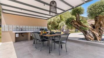 An outdoor dining area with a table and chairs, a hanging light fixture, and a large tree.