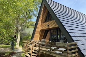 Eine Holzhütte mit geneigtem Dach, eine Holzterrasse mit Sitzgelegenheiten und ein Schild mit Namen und Telefonnummer.