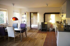 A dining area with a table and chairs, a kitchen with a stove, and a window with curtains. - Winningmay - Magnolia (Schaalby)