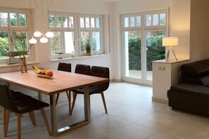 A dining area with a wooden table, black chairs, and a fruit bowl. There are windows with white frames and a view of greenery outside. - Holiday home with 2 bedrooms (Ostseeheilbad Zingst)