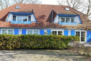 Een tweelaags huis met een rode pannendak, blauwe luiken en een haag op de voorgrond.