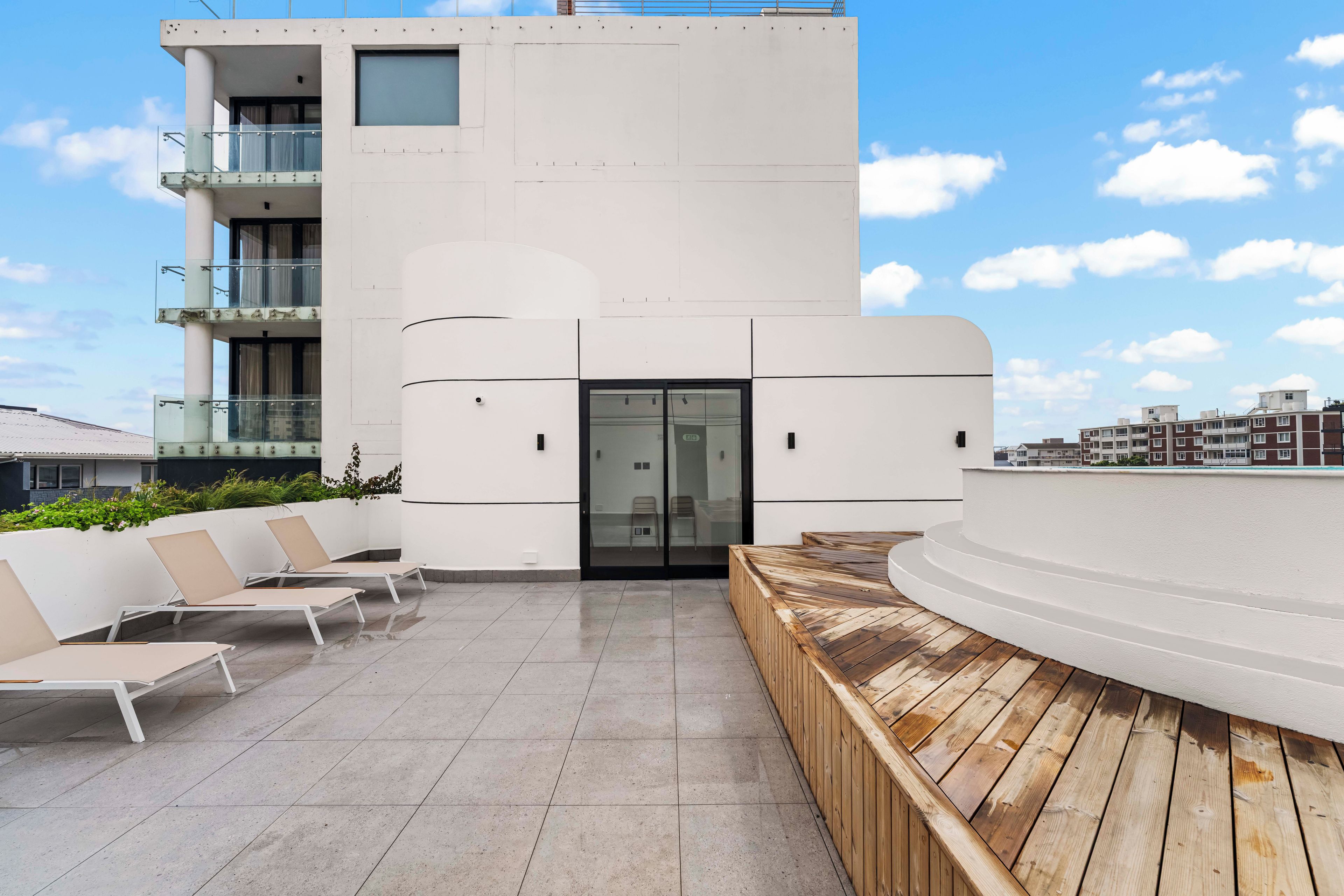 Un edificio moderno con techo plano, puerta de vidrio y un área de estar con sillas y terraza de madera.