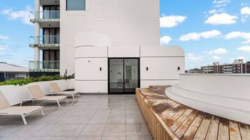 Un edificio moderno con techo plano, puerta de vidrio y un área de estar con sillas y terraza de madera.