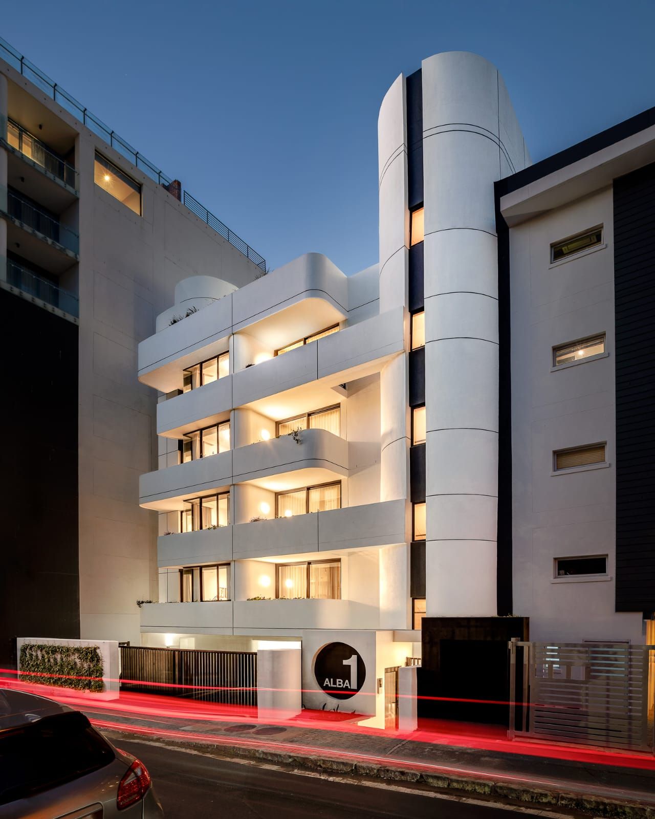 A multi-story hotel with a modern design, featuring balconies on each floor, a prominent cylindrical structure on the right, and a visible entrance with a sign.