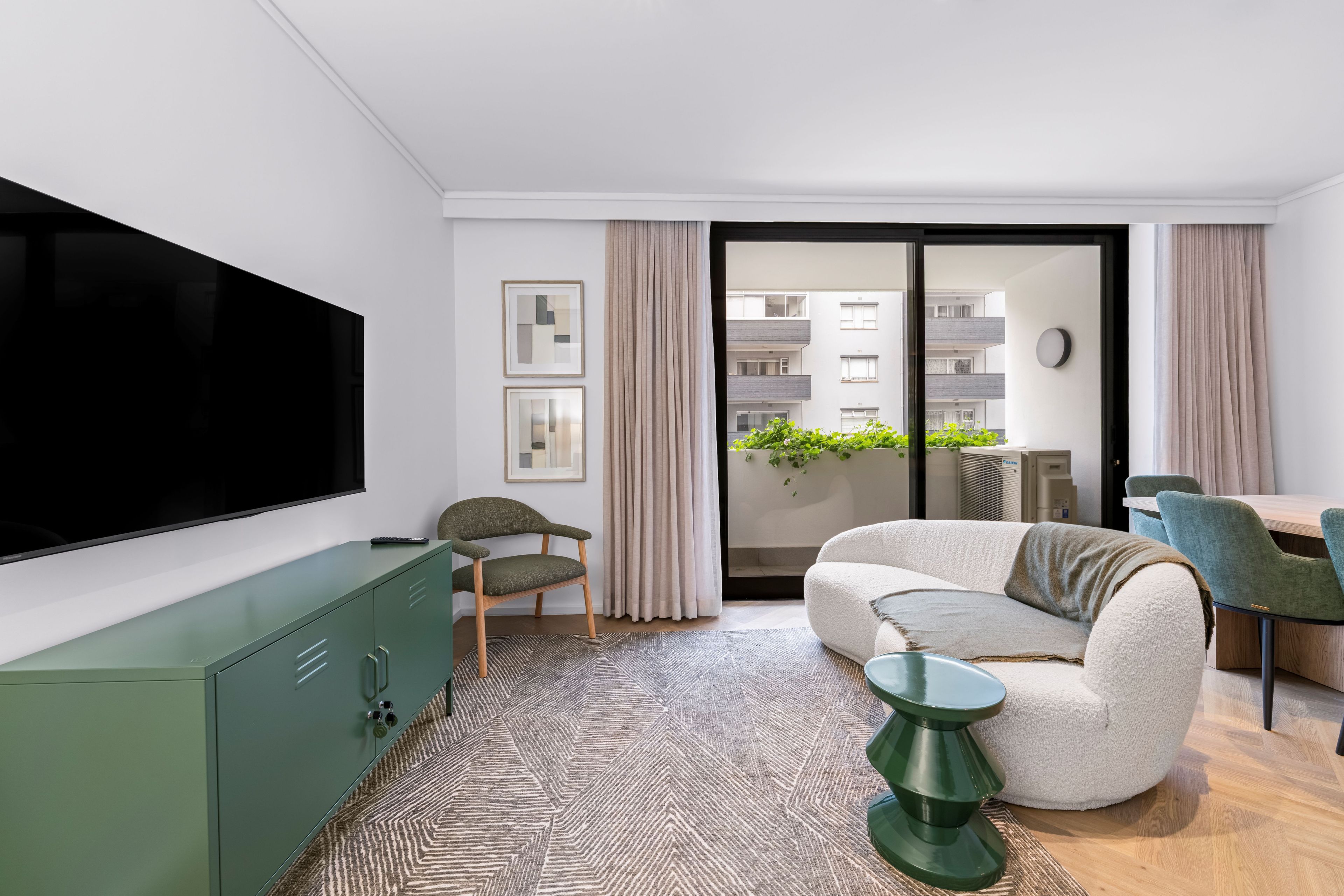 Una sala moderna con una televisión de pantalla plana, un sofá, una mesa de comedor y vistas a un edificio a través de una puerta de cristal.