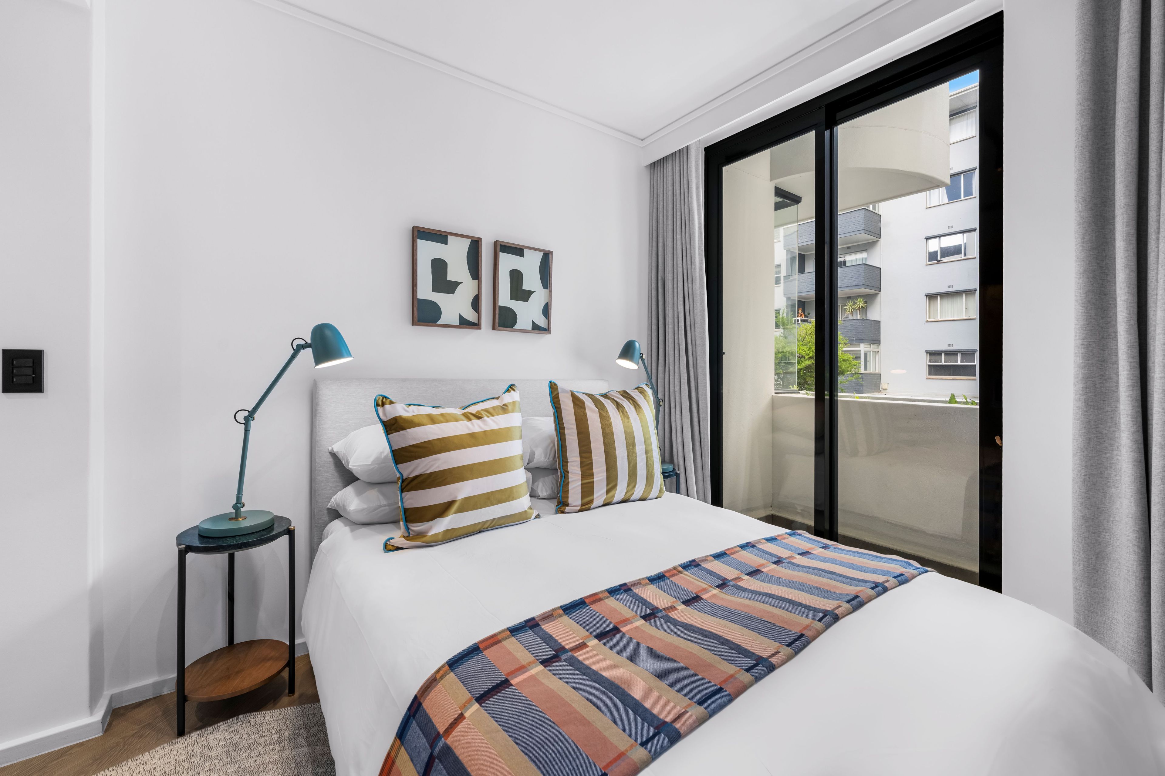A hotel room with a large bed, two bedside lamps, a view of the city through a balcony, and a modern design.