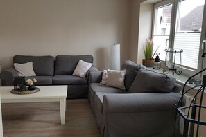Ein Wohnzimmer mit einer grauen Sofagarnitur, einem weißen Couchtisch und einem Fenster mit Blick auf ein Gebäude.