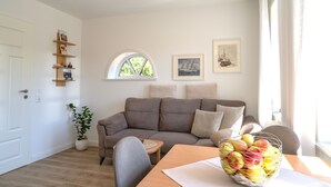 En stue med en sofa, et spisebord med en kurv med frugt, et vindue med udsigt til træer og en væg med billeder i rammer.