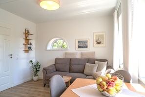 Ein Wohnzimmer mit einer Couch, einem Esstisch mit Obstschale, einem Fenster mit Blick auf Bäume und einer Wand mit gerahmten Bildern.
