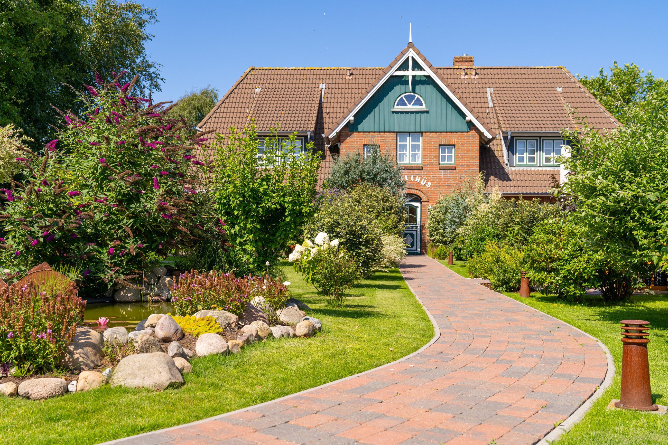 A brick building with a green roof, a pathway leading to the entrance, and a variety of plants and flowers around.