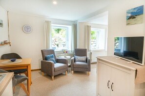 Ein modernes Wohnzimmer mit zwei Sesseln, einem Esstisch, einem Fernseher und Blick auf einen Garten durch die Fenster.