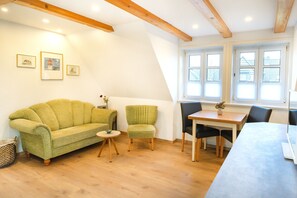 A room with a green sofa, a small round table, a dining table with chairs, and a window with blinds. - Enjoy the cozy Klaas vacation apartment at Trollhüs in Norddorf on Amrum (Norddorf auf Amrum)