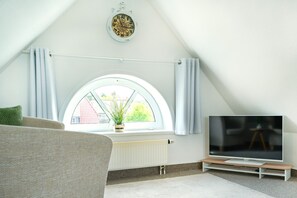 Quarto com teto inclinado, uma janela grande com cortinas, um radiador, uma televisão sobre um suporte e um vaso de planta no parapeito.