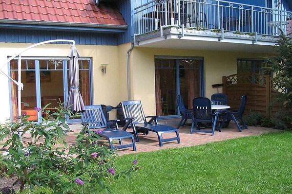 Ein zweistöckiges Gebäude mit einem Balkon, einer Außenbereich-Sitzgelegenheit mit Stühlen und Tischen sowie einem Garten mit Pflanzen und Blumen.
