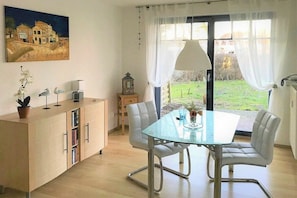 Ein Essbereich mit einem Glastisch, weißen Stühlen, einer Schrank mit Büchern, einem Gemälde an der Wand und Blick auf einen Garten durch eine Tür.
