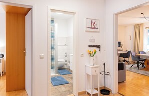 Ein modernes Interieur mit einer Holztür, einem Badezimmer mit Glasdusche, einem Essbereich mit Tisch und Stühlen sowie einer Vase mit Blumen auf einem kleinen weißen Beistelltisch.