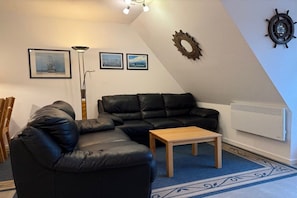 A living room with a black leather sofa, a wooden coffee table, and a radiator. There are framed pictures on the wall and a floor lamp. - Holiday apartment with 2 bedrooms (Büsumer Deichhausen)