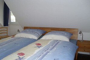 A bedroom with a sloped ceiling, a wooden headboard, two beds with blue and white bedding, a nightstand with a lamp, and a window with curtains. - Holiday home with 3 bedrooms (St.Peter-Ording)