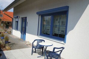 A building with a white exterior and blue trim, featuring a window with a view of a house, a door, and a small outdoor seating area with two chairs and a table. - Holiday home with 1 bedroom (Insel Poel - OT Kaltenhof)