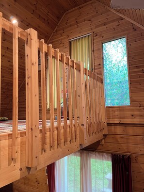 Un intérieur en bois avec un balcon, une fenêtre donnant sur un paysage verdoyant et un plafond avec des poutres apparentes.