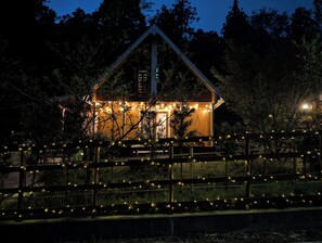 A house with a triangular roof, surrounded by trees and a wooden fence with string lights.
